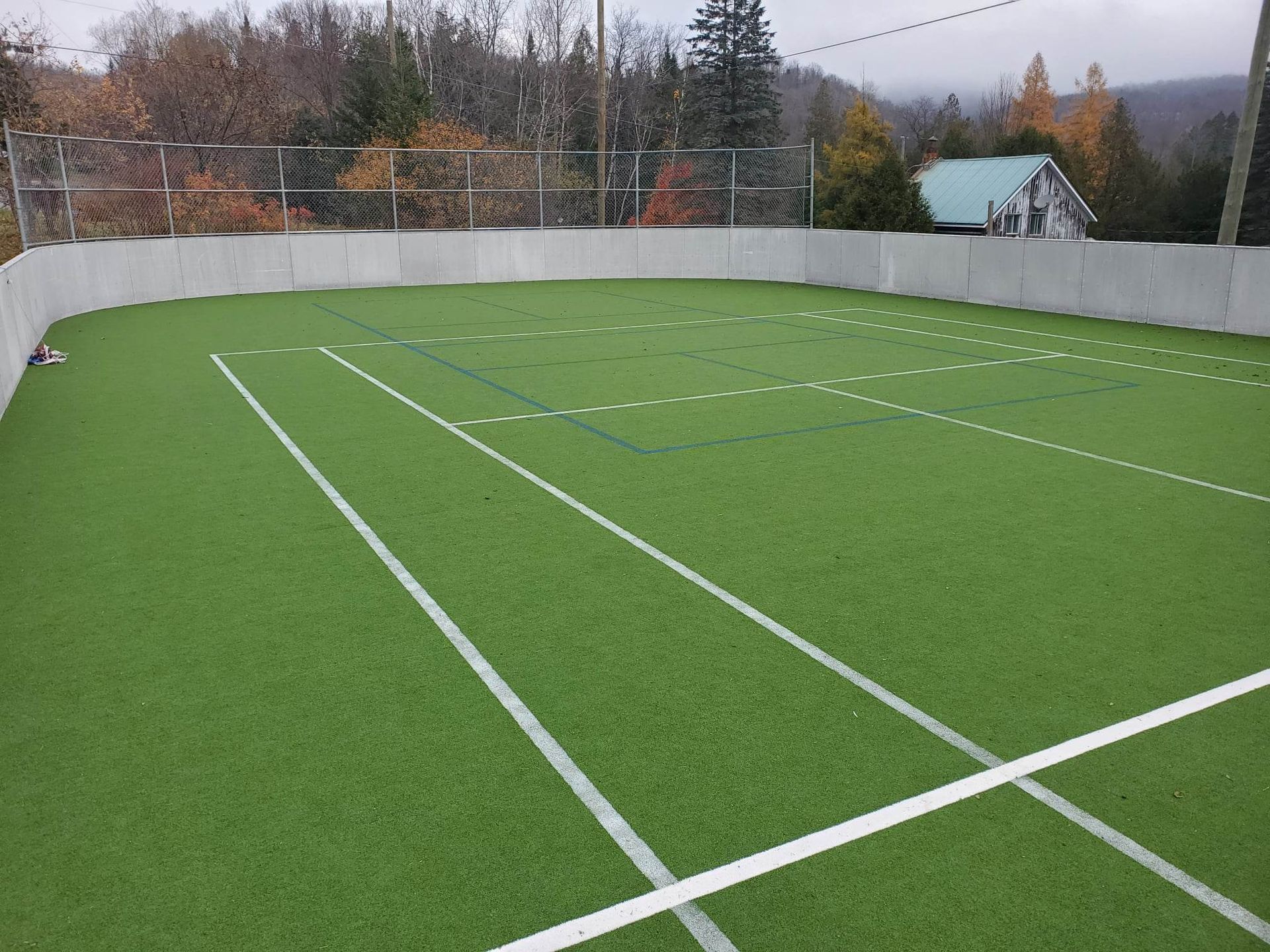 Terrain de sport en gazon artificiel avec des lignes blanches et un mur gris, sur fond d'arbres et d'une maison sous un ciel couvert.