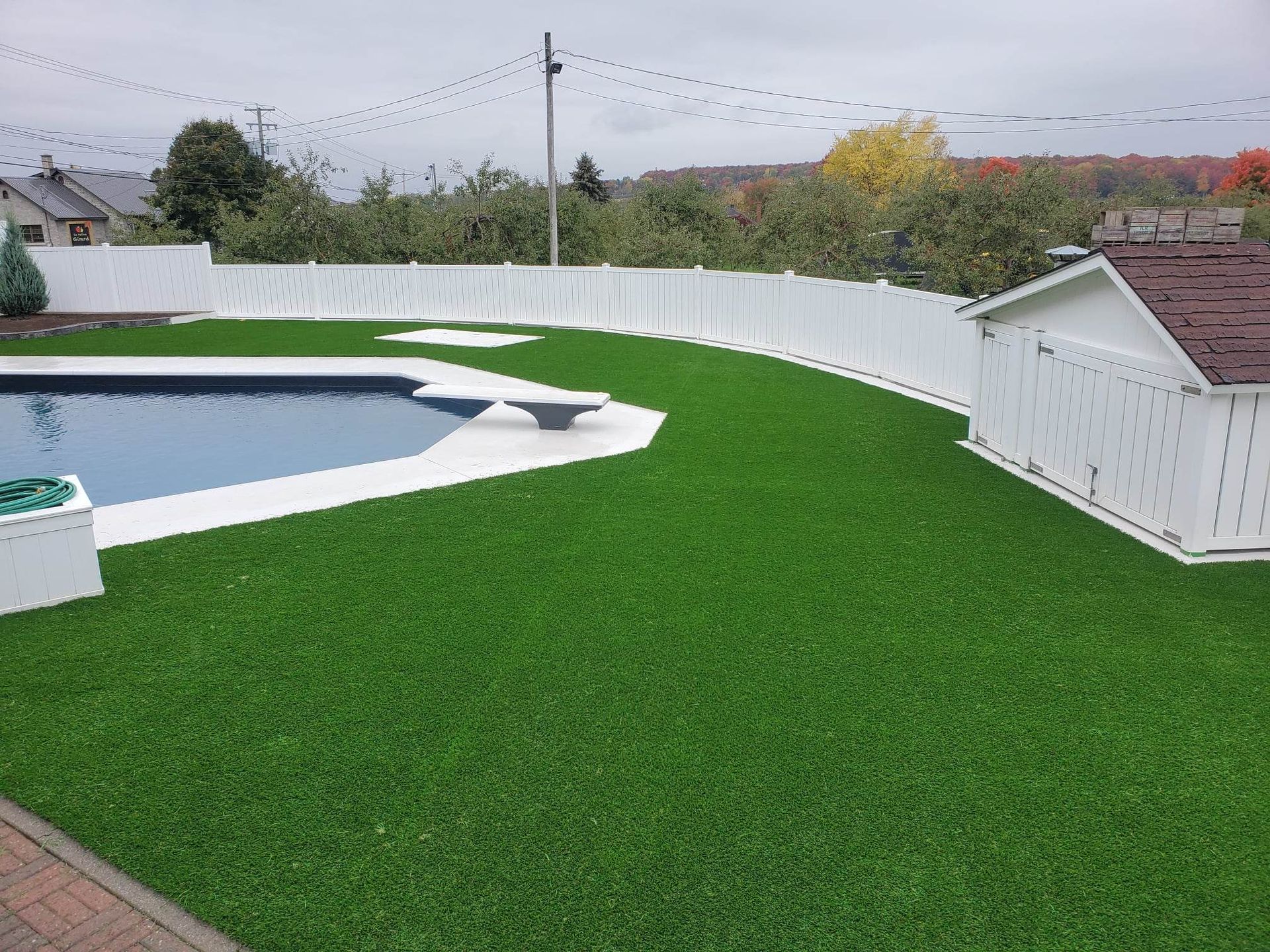 Un jardin avec piscine, entouré d'une clôture blanche et d'un gazon artificiel vert vif. Le ciel est couvert.