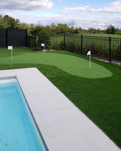 Un jardin avec un putting green, une piscine et des clôtures noires. Trois drapeaux blancs marquent les trous sur le green.