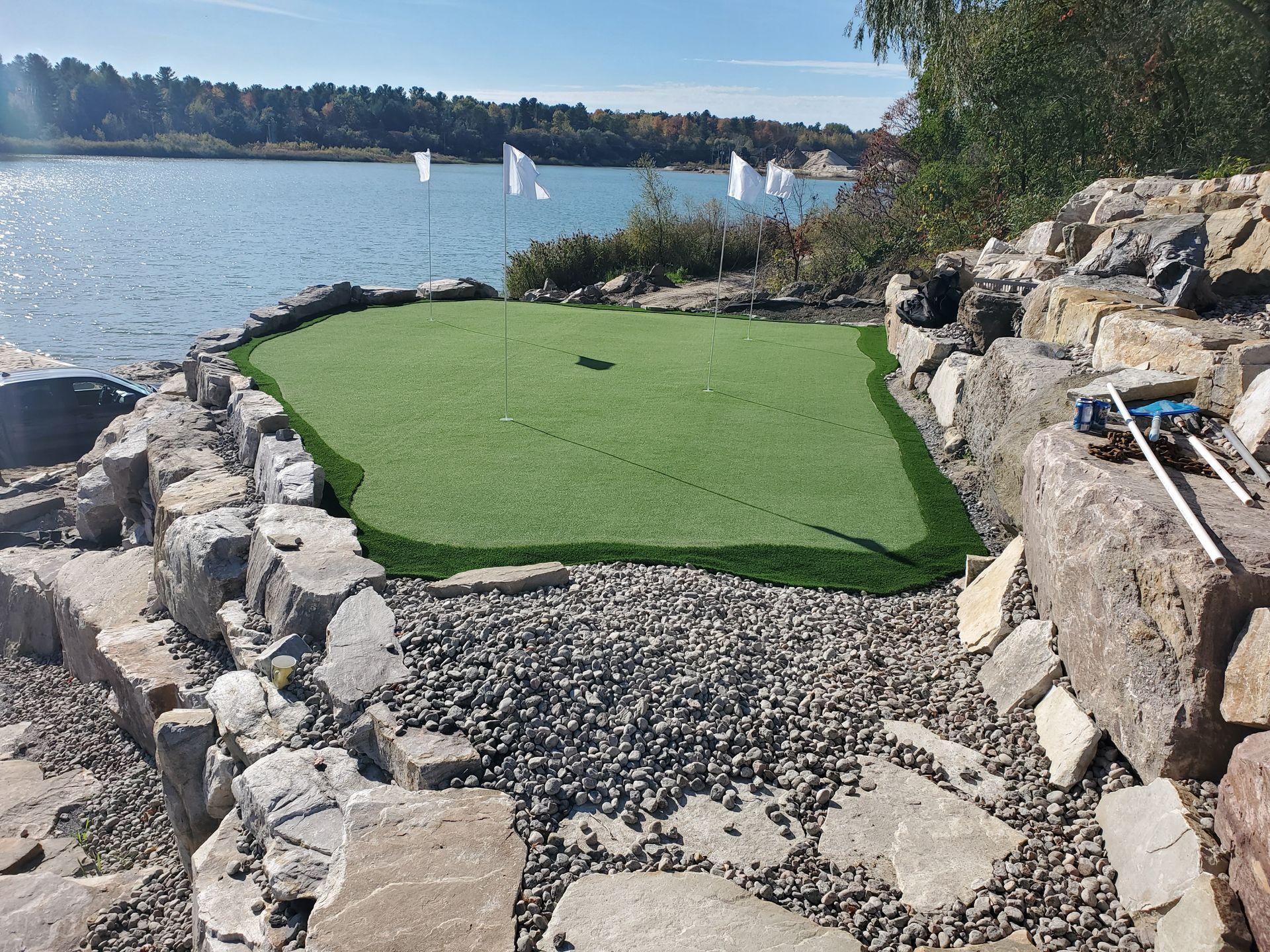Un petit putting green sur une rive rocheuse au bord d'un lac, avec des drapeaux blancs et un ciel bleu.