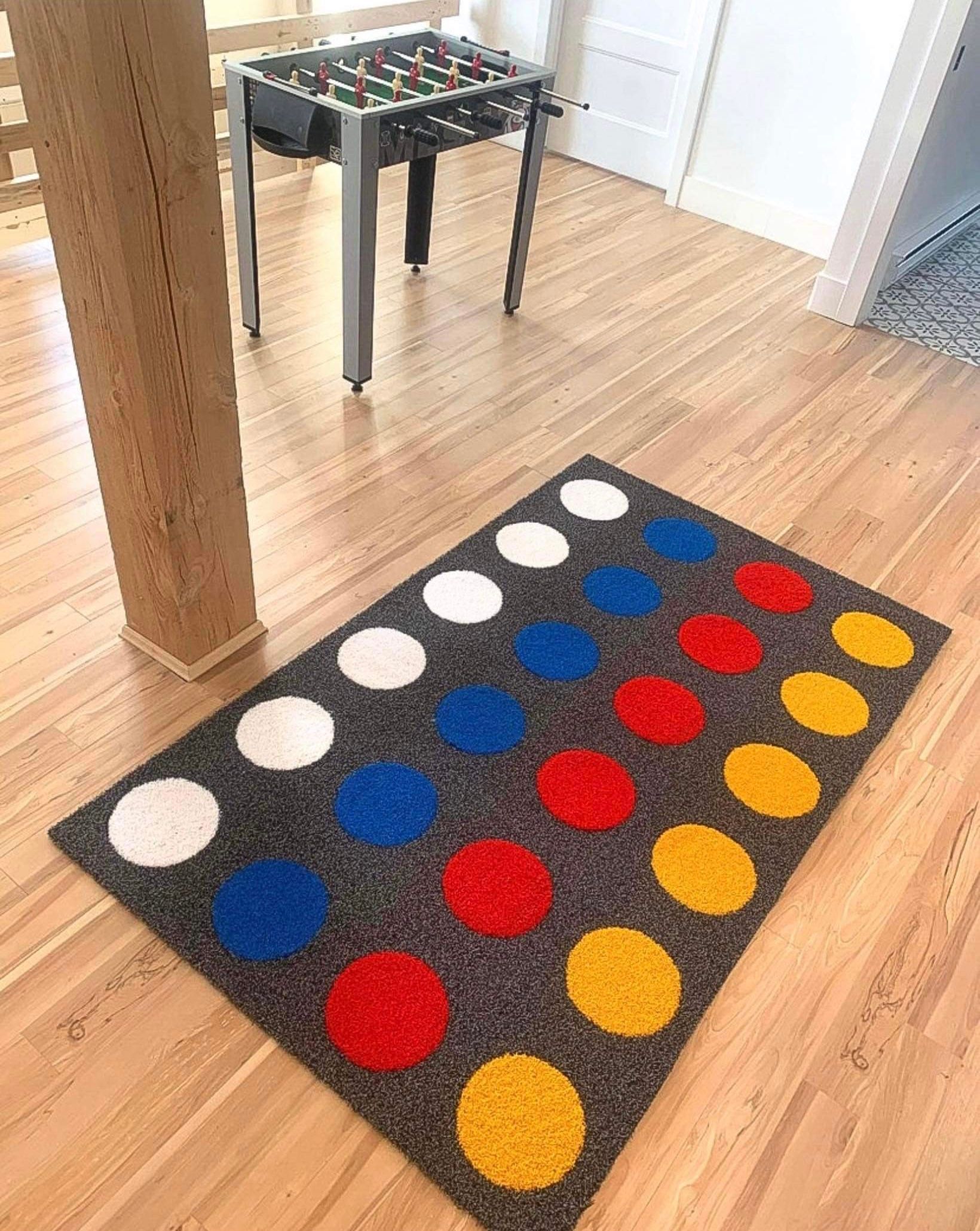 Tapis Twister sur un parquet en bois orné de cercles blancs, bleus, rouges et jaunes. Une petite table de jeu et une colonne texturée sont également présentes.
