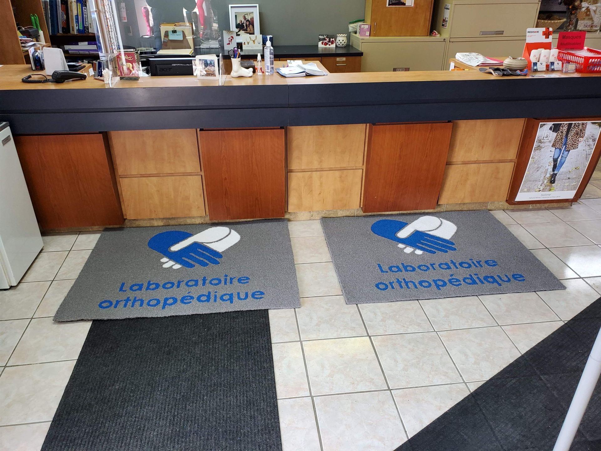Deux tapis gris avec un logo bleu et blanc indiquant « Laboratoire Orthopédique » devant un comptoir en bois.