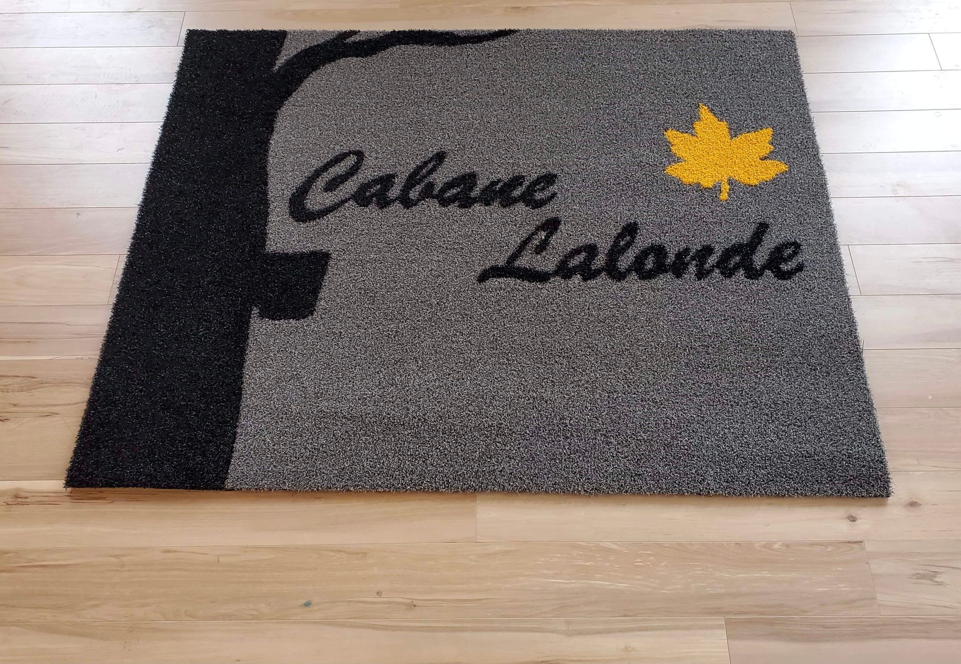 Paillasson gris avec le texte « Cabane Lalonde », une silhouette d'arbre et une feuille jaune sur un plancher en bois.