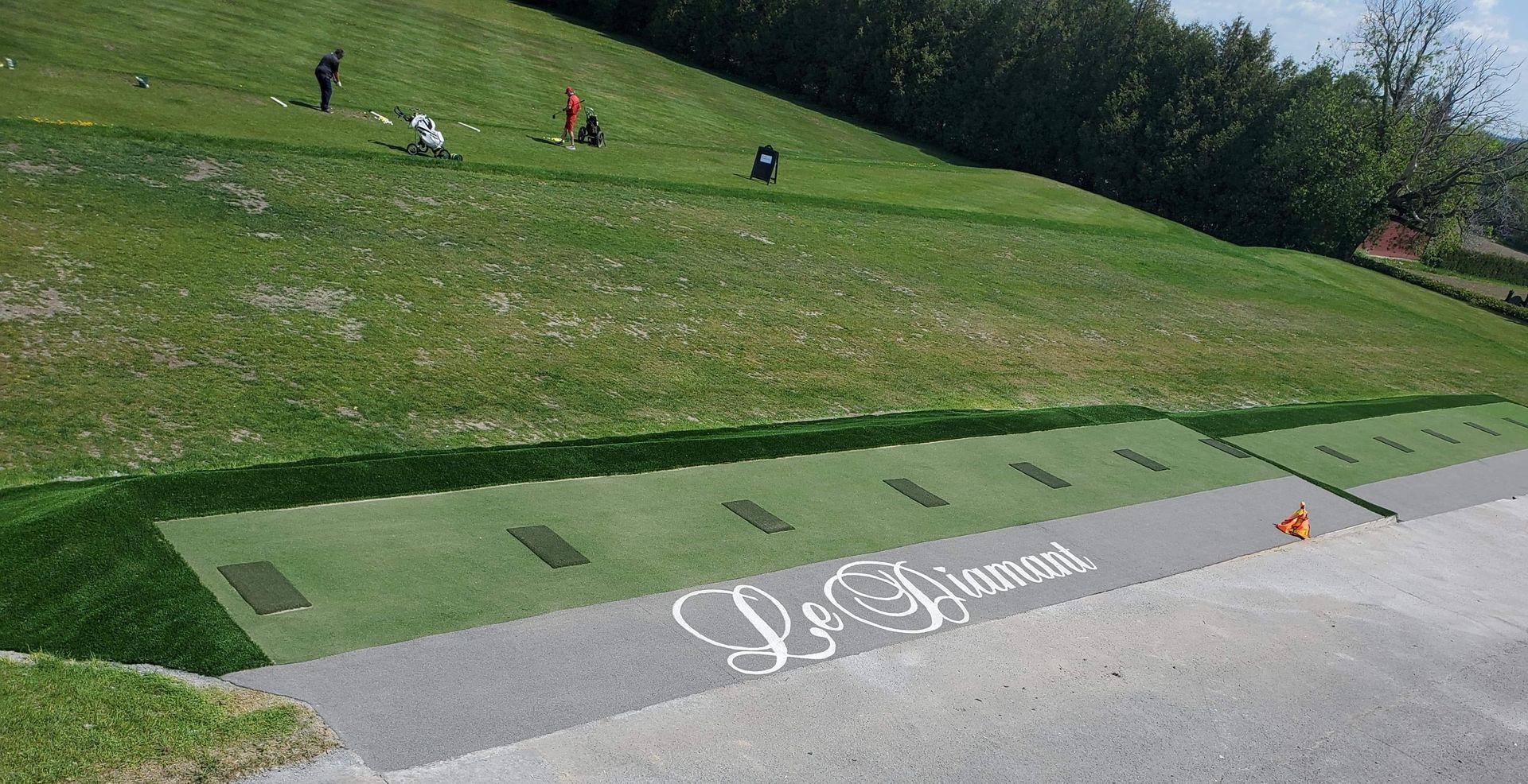 Practice de golf avec des golfeurs sur une colline herbeuse ; départs sur une piste asphaltée. Le logo « La Fleur » est apposé sur l'asphalte.