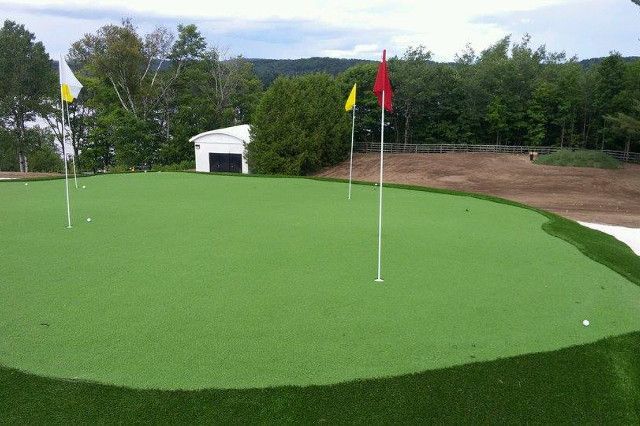Golf vert avec des mâts de drapeau blancs et des drapeaux colorés sur fond d'arbres et d'un petit bâtiment sous un ciel nuageux.