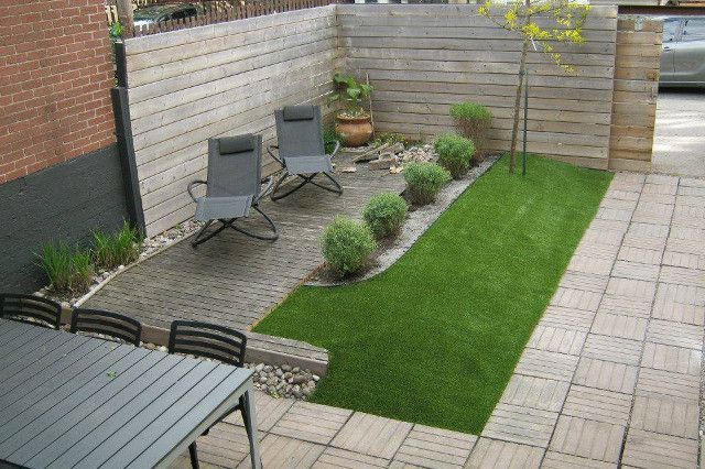 A small urban patio with gray paving stones, artificial turf, wooden fence, and two chairs; a dining table sits in the foreground.