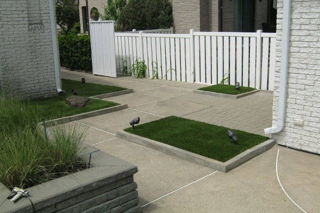 Terrasse en béton avec pelouses rectangulaires. Une clôture blanche et un bâtiment en arrière-plan.