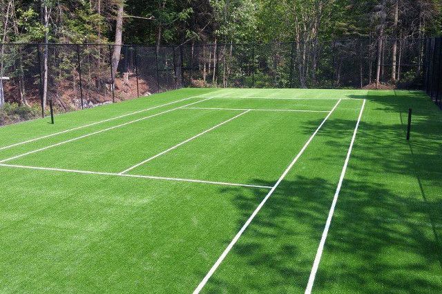 Terrain de tennis avec gazon artificiel vert et lignes blanches, situé dans une zone boisée.