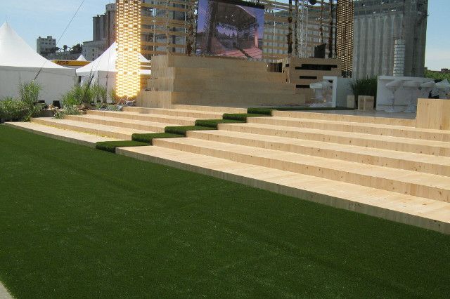 Une scène extérieure avec des gradins, du gazon vert devant et des bâtiments en arrière-plan. Des pierres claires ont été utilisées pour les sièges.