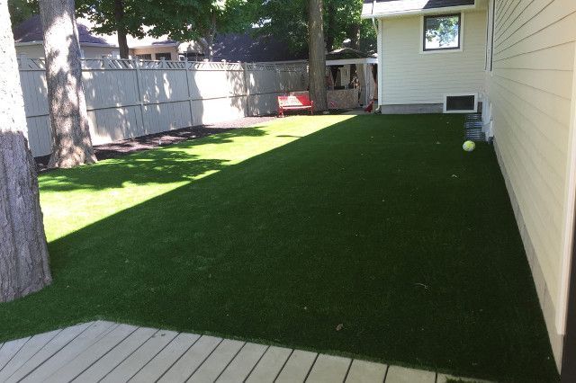 Un jardin avec gazon synthétique vert à côté d'une maison beige. Une terrasse en bois est au premier plan et une clôture blanche au loin.