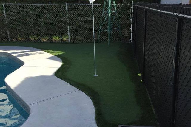 Putting green dans le jardin à côté d'une piscine, avec une clôture noire et une clôture grillagée en arrière-plan.