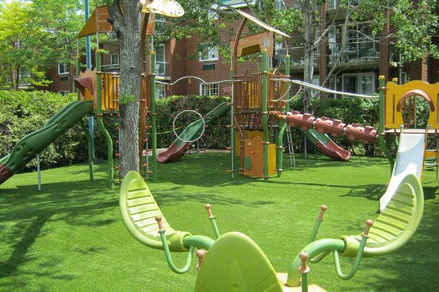 Aire de jeux avec structures vertes et brunes, dont des toboggans et une balançoire, sur gazon vert, avec un bâtiment en briques en arrière-plan.