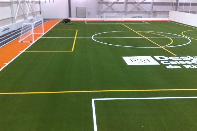 Terrain de football intérieur avec gazon vert, lignes blanches et jaunes et but. Bordure orange et grand logo visibles.