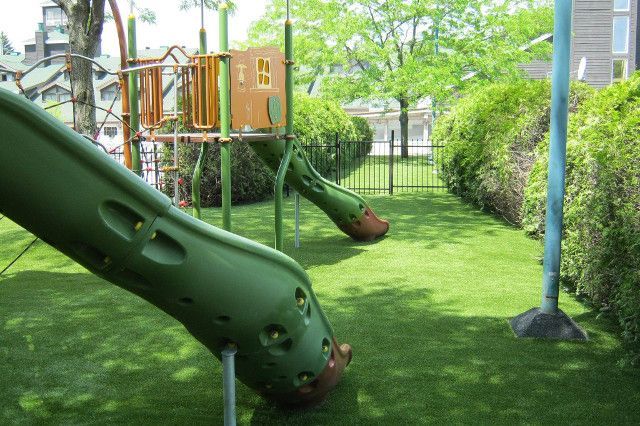 Une aire de jeux verdoyante avec toboggan, installée sur du gazon synthétique. Une clôture et des buissons verts entourent l'aire de jeux.