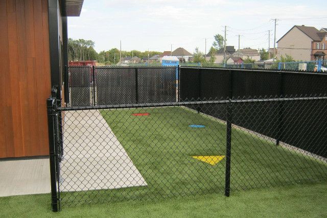 Une clôture grillagée noire entoure une zone de gazon artificiel vert avec des marqueurs colorés, à côté d'un bâtiment en bois.