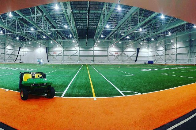 Terrain de sport intérieur avec gazon vert, piste orange et véhicule utilitaire. Un éclairage zénithal puissant illumine ce vaste espace.