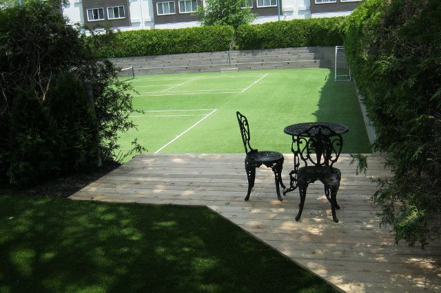 Terrasse en bois avec table et chaises décorées donnant sur un court de tennis vert, entouré de haies, une journée ensoleillée.