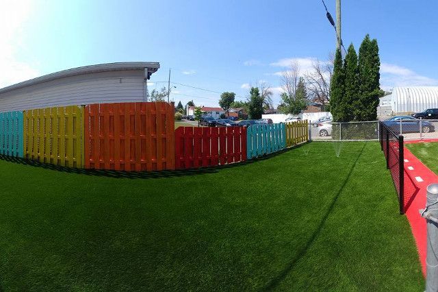 Clôture colorée dans une cour verte ; bâtiments et voitures en arrière-plan sous un ciel bleu.