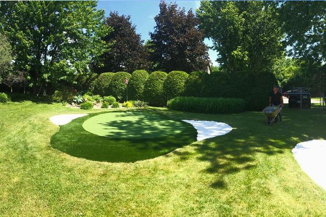 Un putting green dans l'arrière-cour entouré d'une bordure blanche, d'une herbe verte luxuriante et d'un aménagement paysager ; une personne travaille à droite.