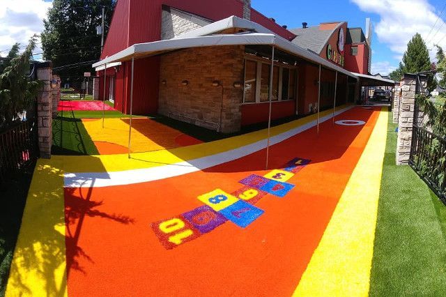 Une aire de jeux extérieure colorée avec un parcours de marelle devant un bâtiment rouge.