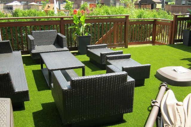 Outdoor patio with black wicker furniture on green artificial turf, enclosed by a wooden railing and green hedges.