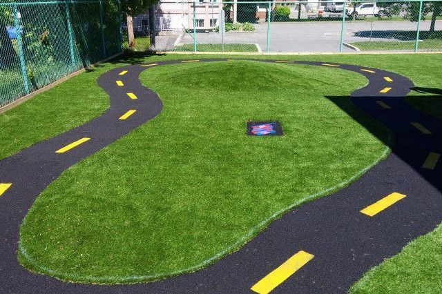 A child's play area with black asphalt roads and yellow lane markers winding through green artificial turf, surrounding a small hill.