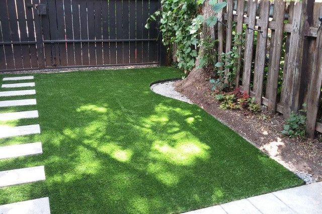 Terrain en gazon synthétique avec pas japonais, bordé d'une clôture en bois et d'un tronc d'arbre, avec ombre.