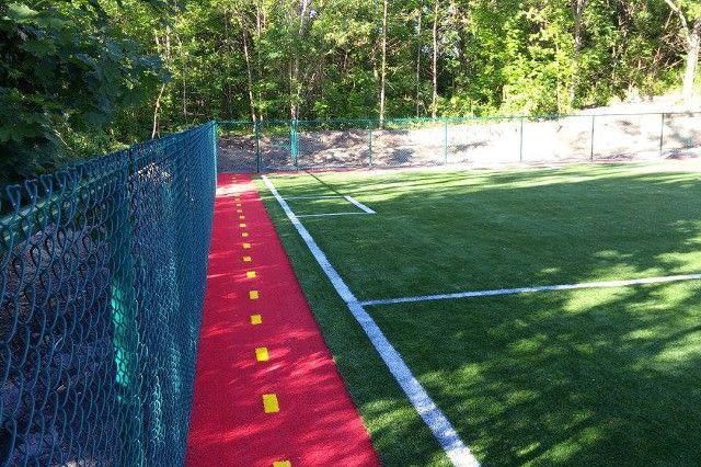 Piste de course rouge avec lignes pointillées jaunes à côté d'un terrain en gazon synthétique vert, séparé par une clôture grillagée. Des arbres apparaissent en arrière-plan.
