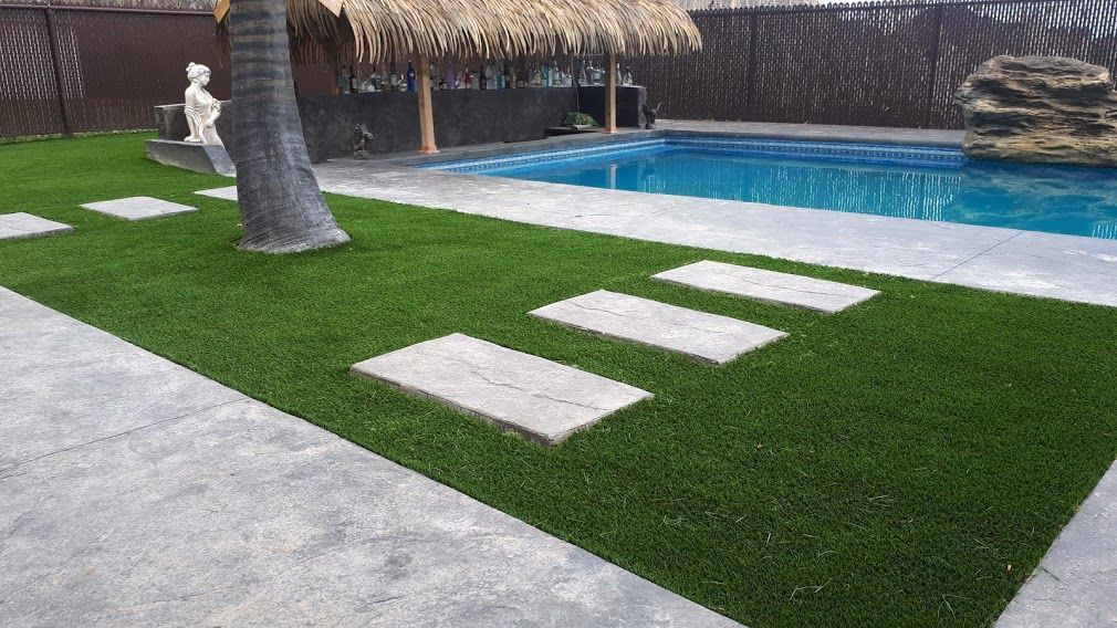 Cour en gazon artificiel vert avec des dalles en béton, une piscine et une structure au toit de chaume.