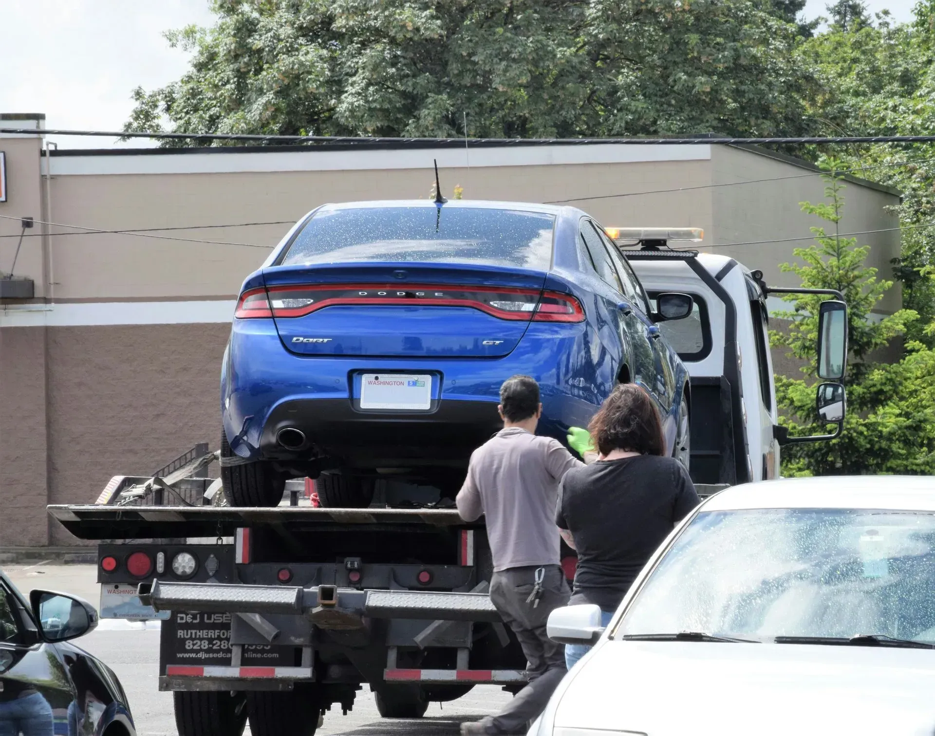 Blue car being loaded onto a tow truck while two people stand nearby outside a building