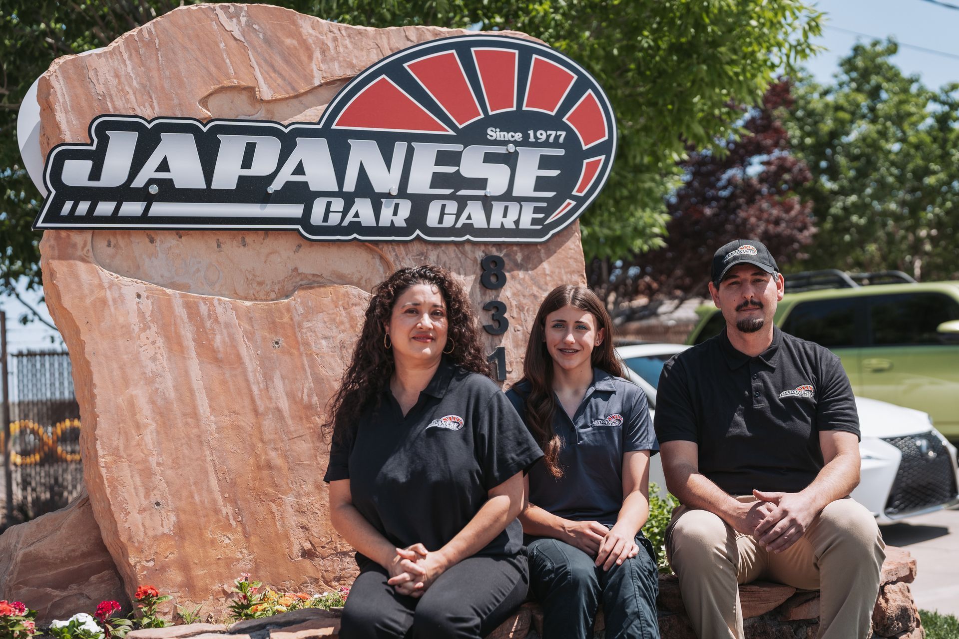 Three people are posing for a picture in front of a japanese car care sign. | Japanese Car Care