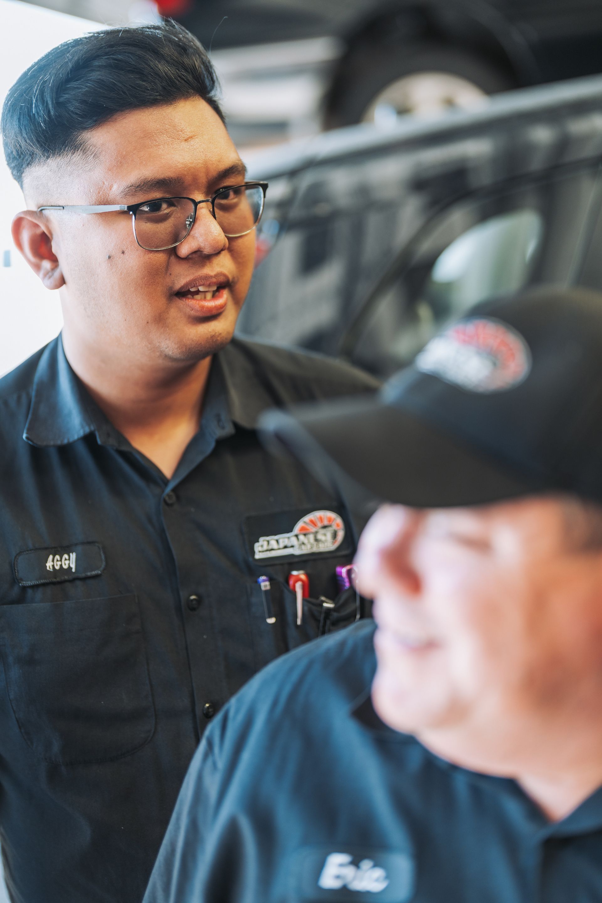 Two men are standing next to each other in a garage talking to each other. | Japanese Car Care