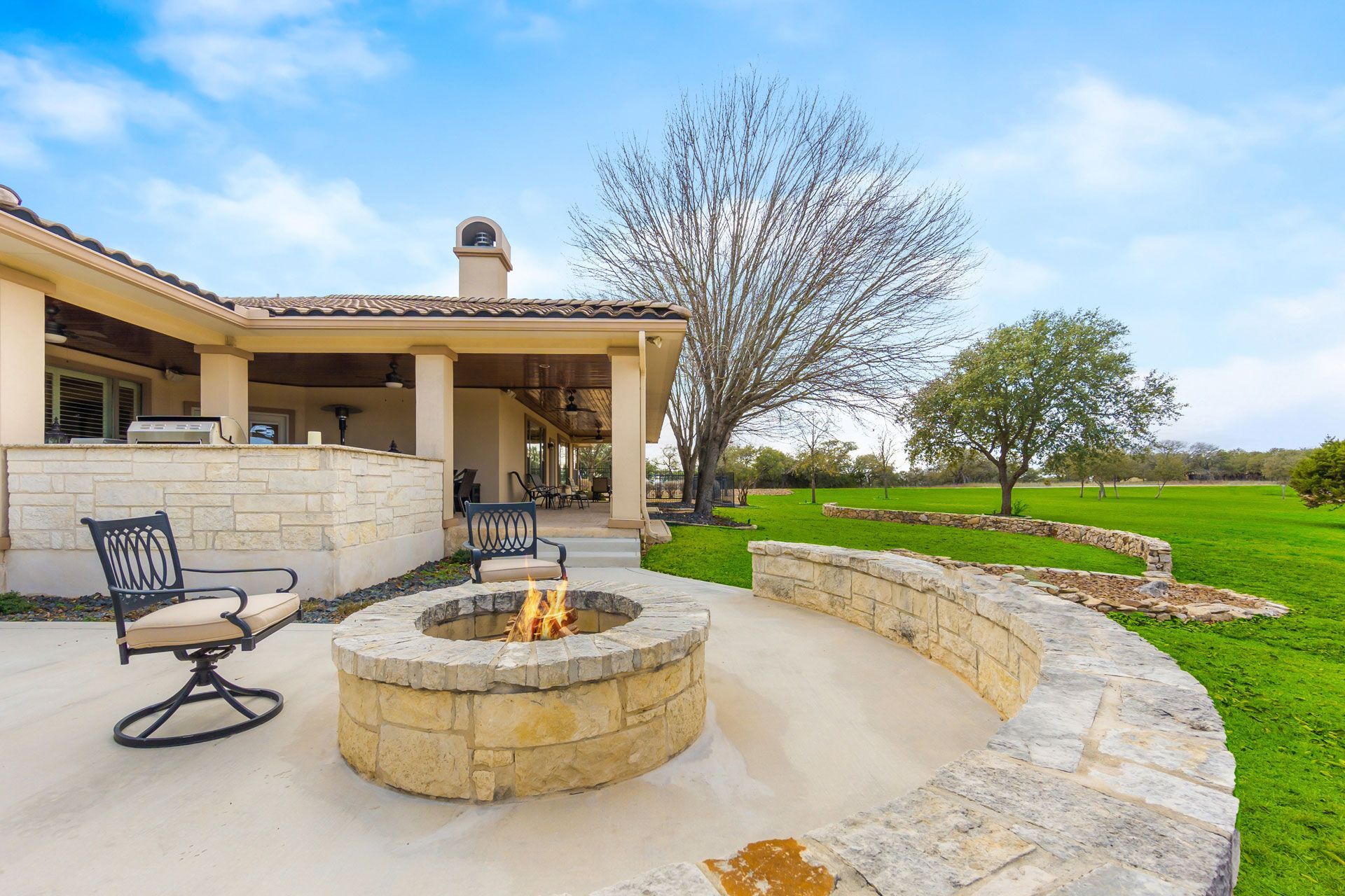 Patio with stone fire pit, lounge chairs, and curved wall beside a house and green lawn under a blue sky