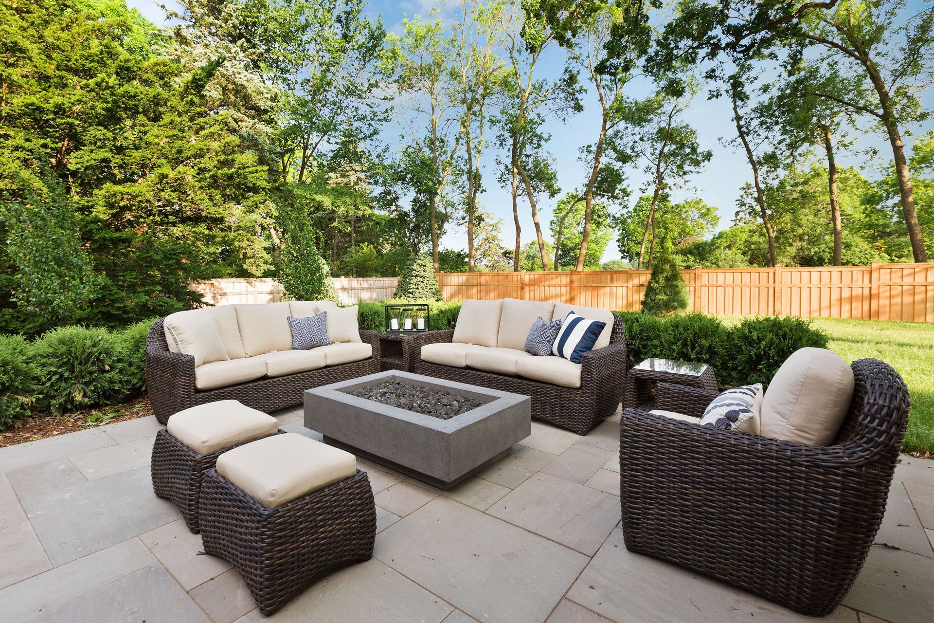 Outdoor patio seating set with dark wicker sofas, beige cushions, and a fire pit on a stone patio.