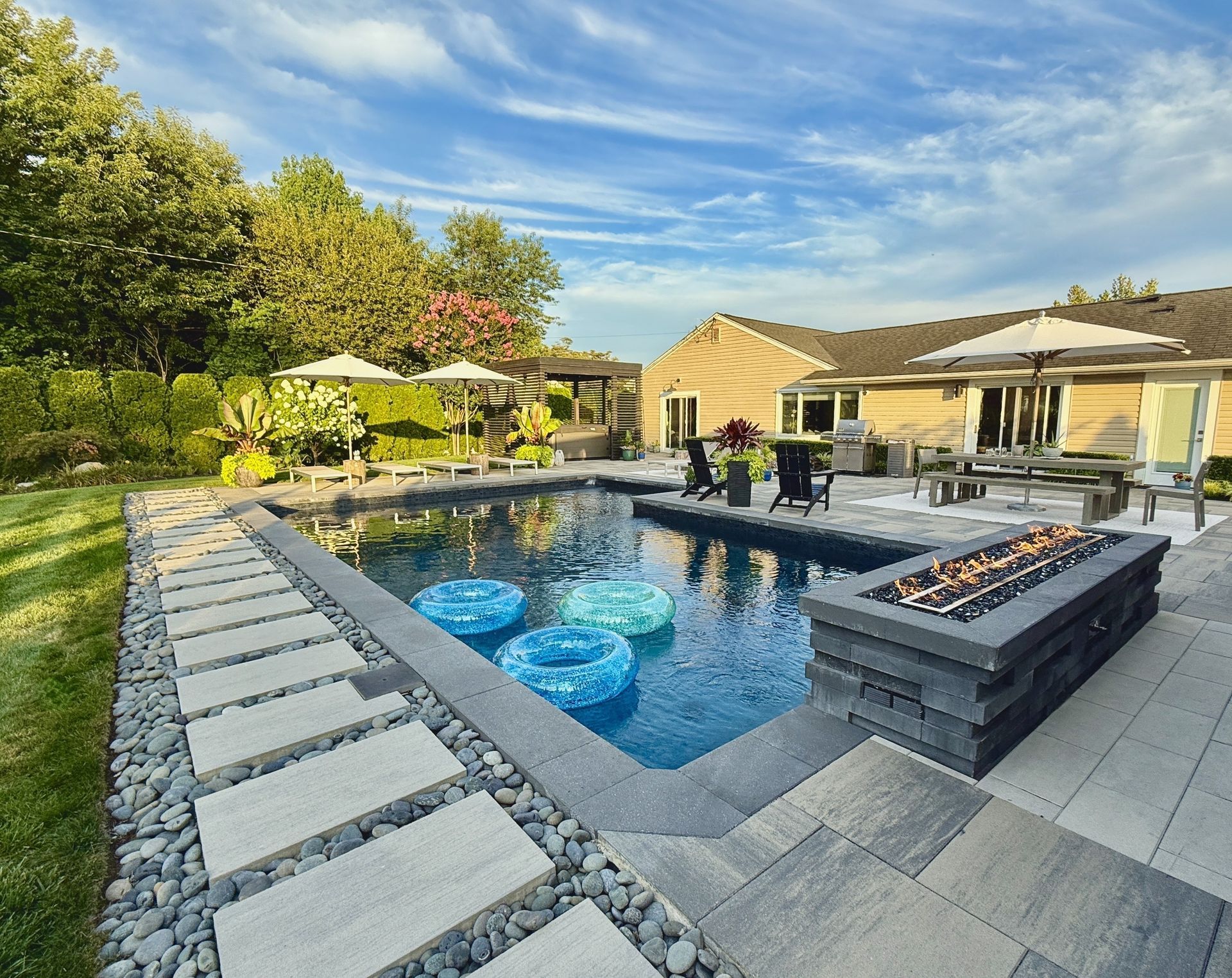 A rectangular backyard pool with three floating tubes, a stone fire pit, and a dining area beside a tan-sided house.