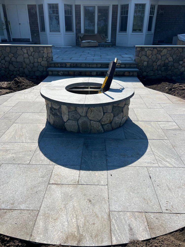 A stone circular fire pit sits in the center of a gray patio in front of a house, with a dark cover partially open.