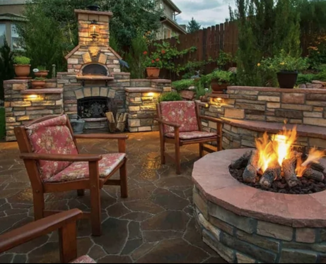An outdoor stone patio with a fire pit, a built-in stone fireplace with an oven, and wooden armchairs with floral cushions.