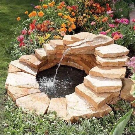 A tiered stone fountain flows into a small, circular pool, surrounded by vibrant flowers and lush greenery.