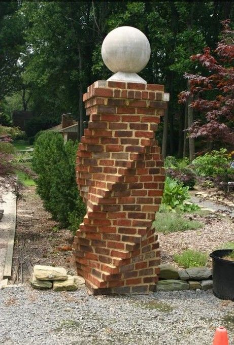 A decorative red brick pillar with a twisted design, topped with a stone sphere, standing in a landscaped garden.