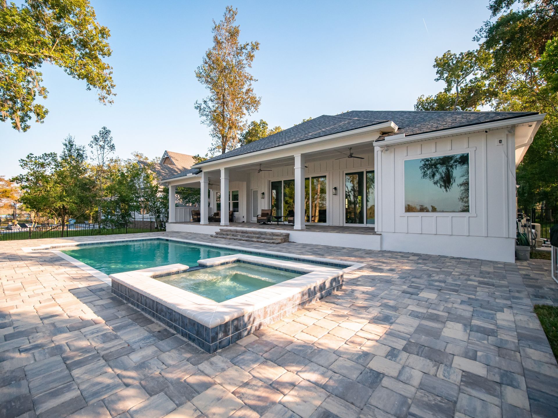 A white house with a covered patio overlooks a backyard swimming pool and hot tub surrounded by stone pavers.