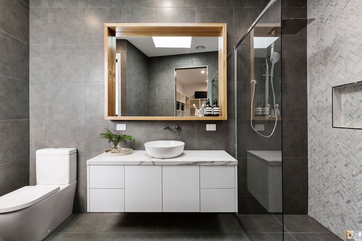 Grey bathroom with white toilet, white under sink cabinets and mirror with wooden frame.