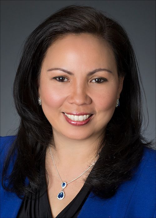 Woman with dark hair smiles, wearing a blue blazer and pendant necklace against a gray backdrop.