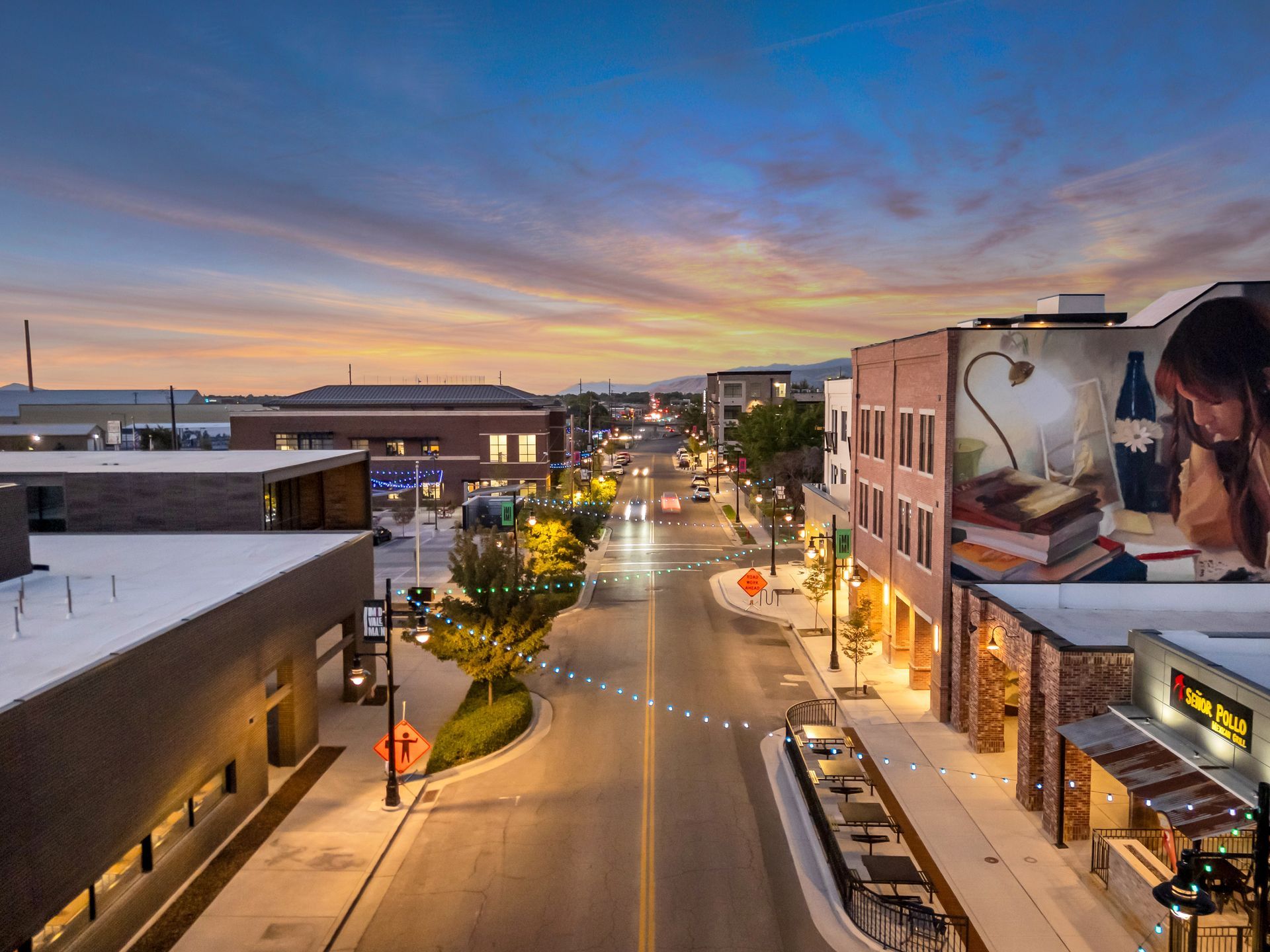 Street at dusk in a town, with illuminated buildings and colorful sky, a mural on one building.