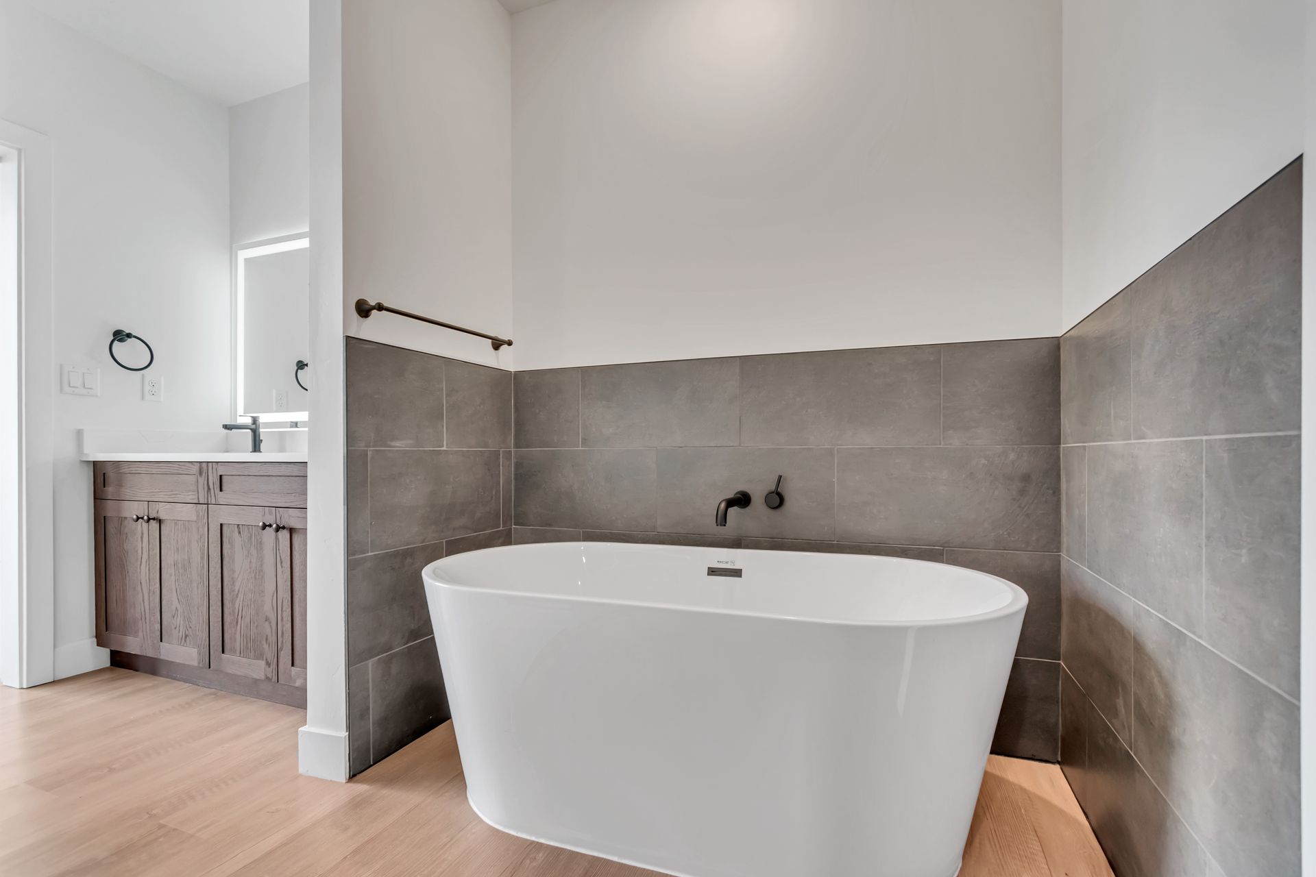 Bathroom with white tub, gray tiled wall, and wood cabinets.
