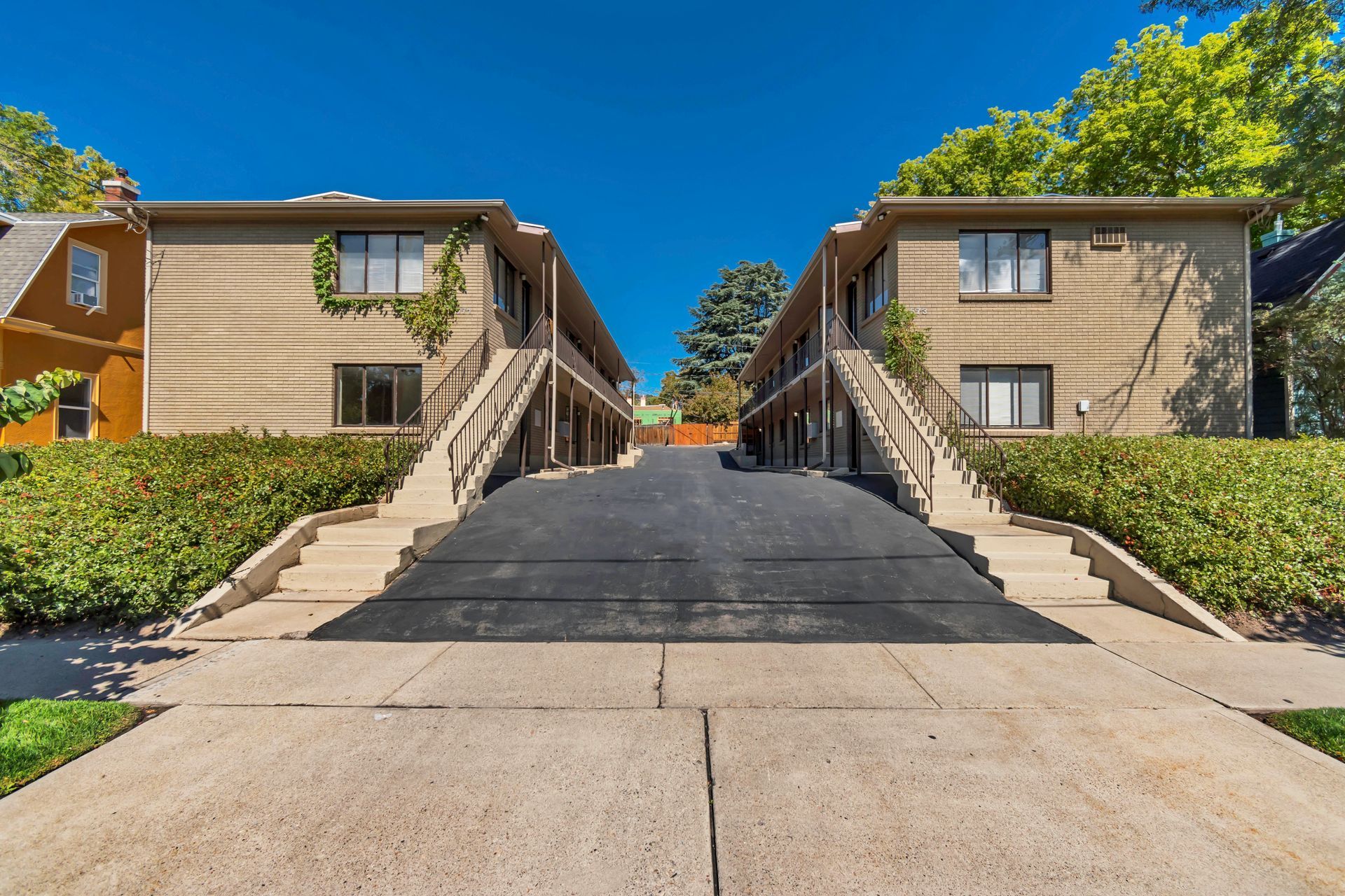 Two-story tan apartment buildings face each other, connected by a paved driveway. Lush green bushes and blue sky.