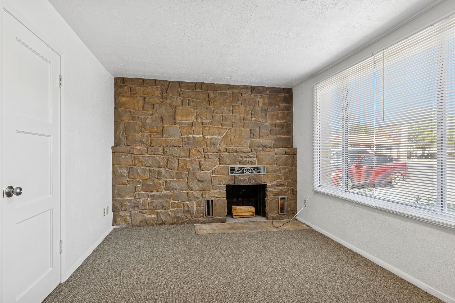 Empty room with stone fireplace, carpet, window with blinds, and a white door.