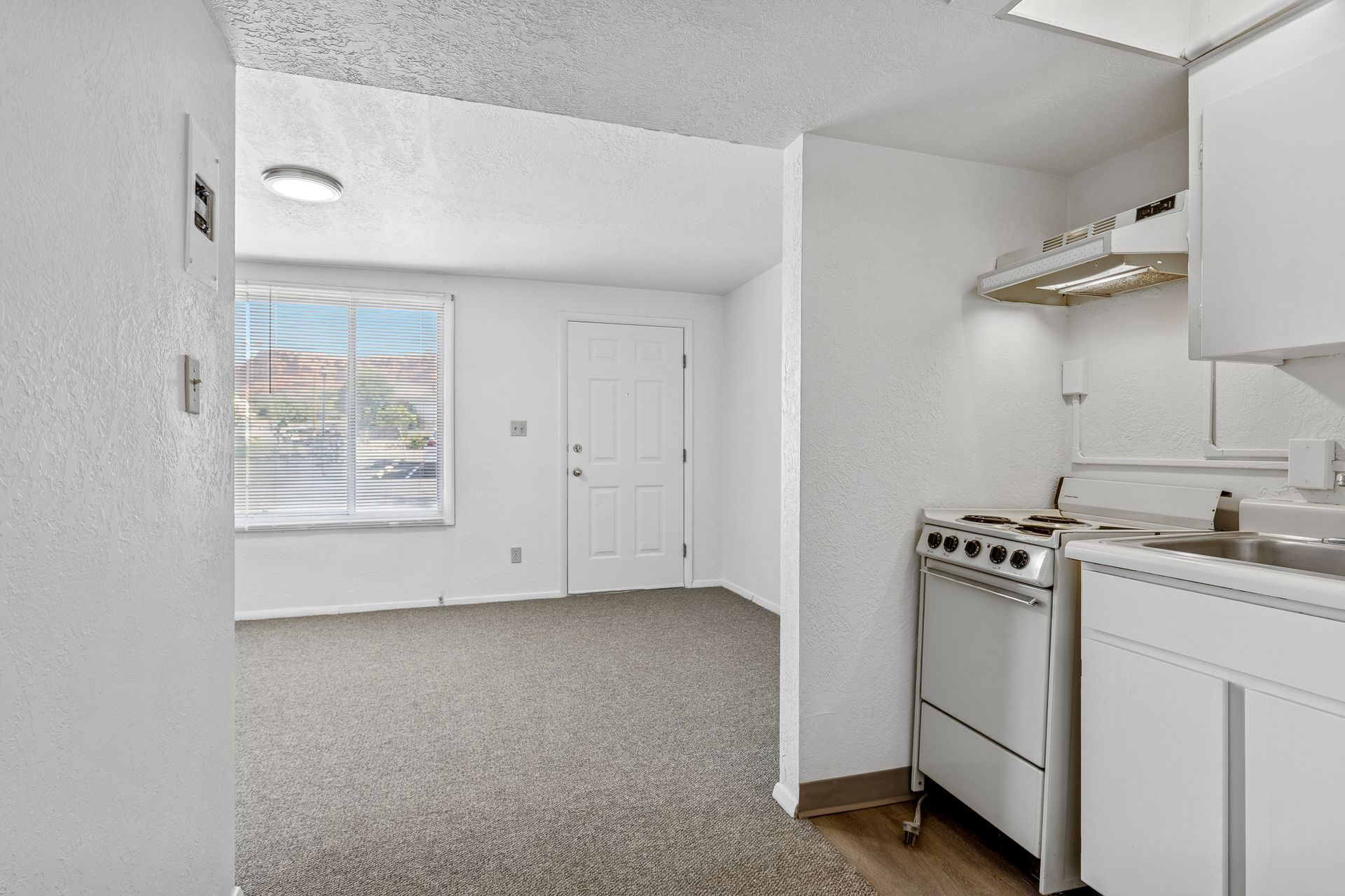 A small, white kitchen with a stove and a view into a living area with a window and a door.