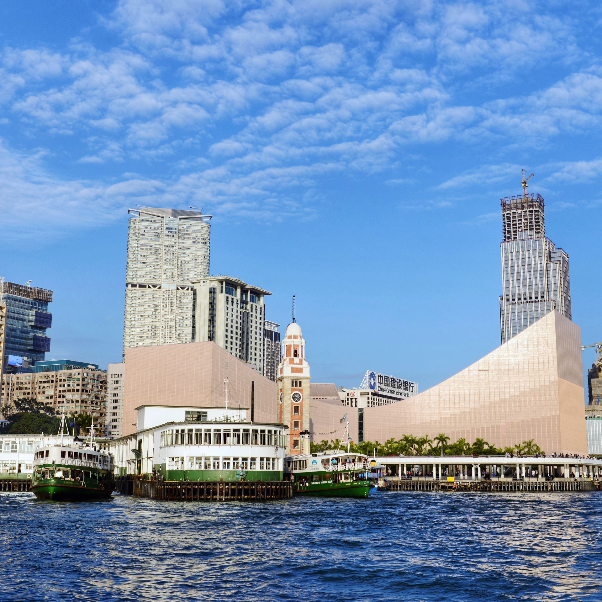 Hong Kong skyline with blue water, ferries, and modern buildings under a cloudy sky.