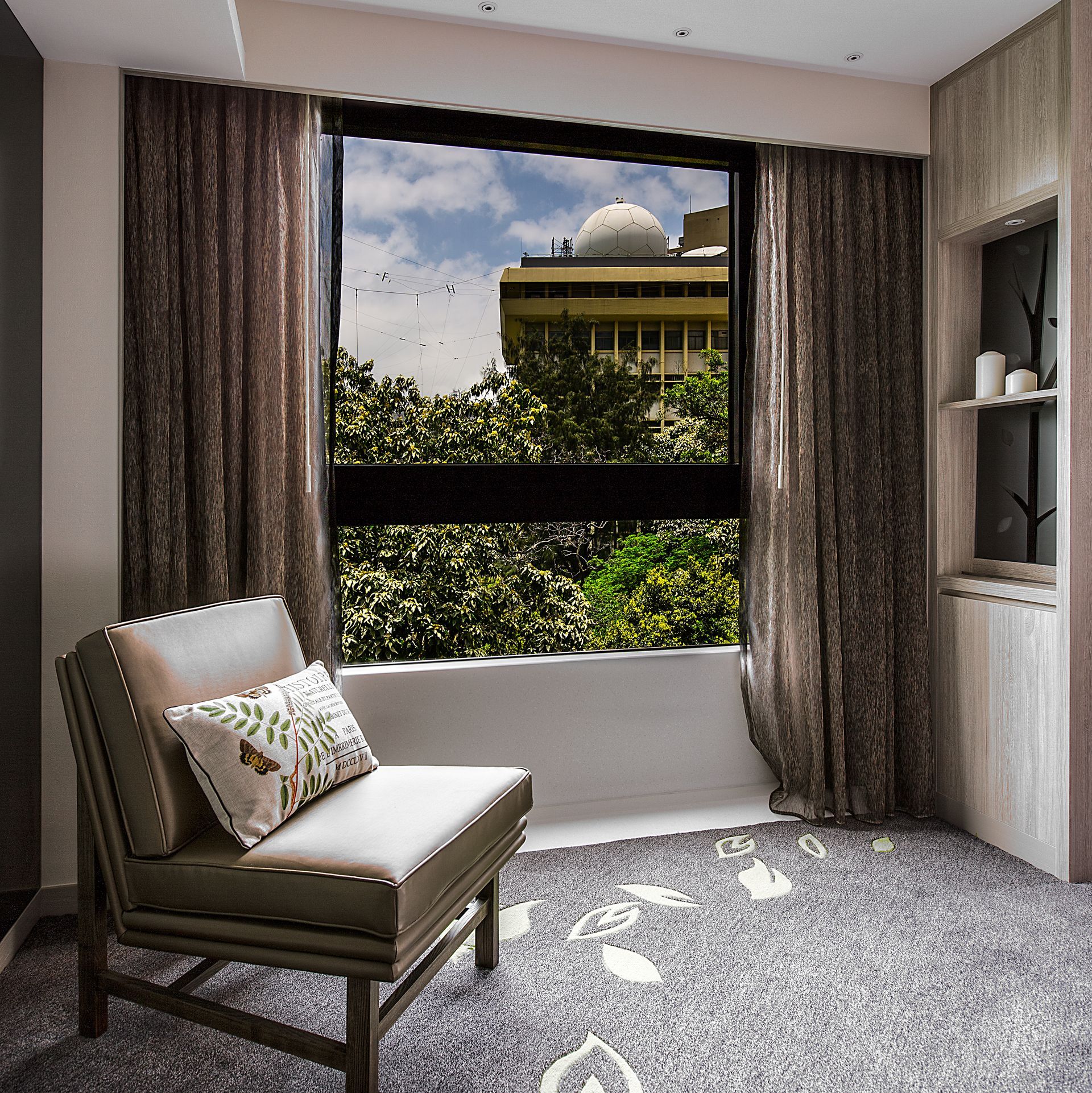 Chair by window with view of trees and building, brown curtains, gray carpet.