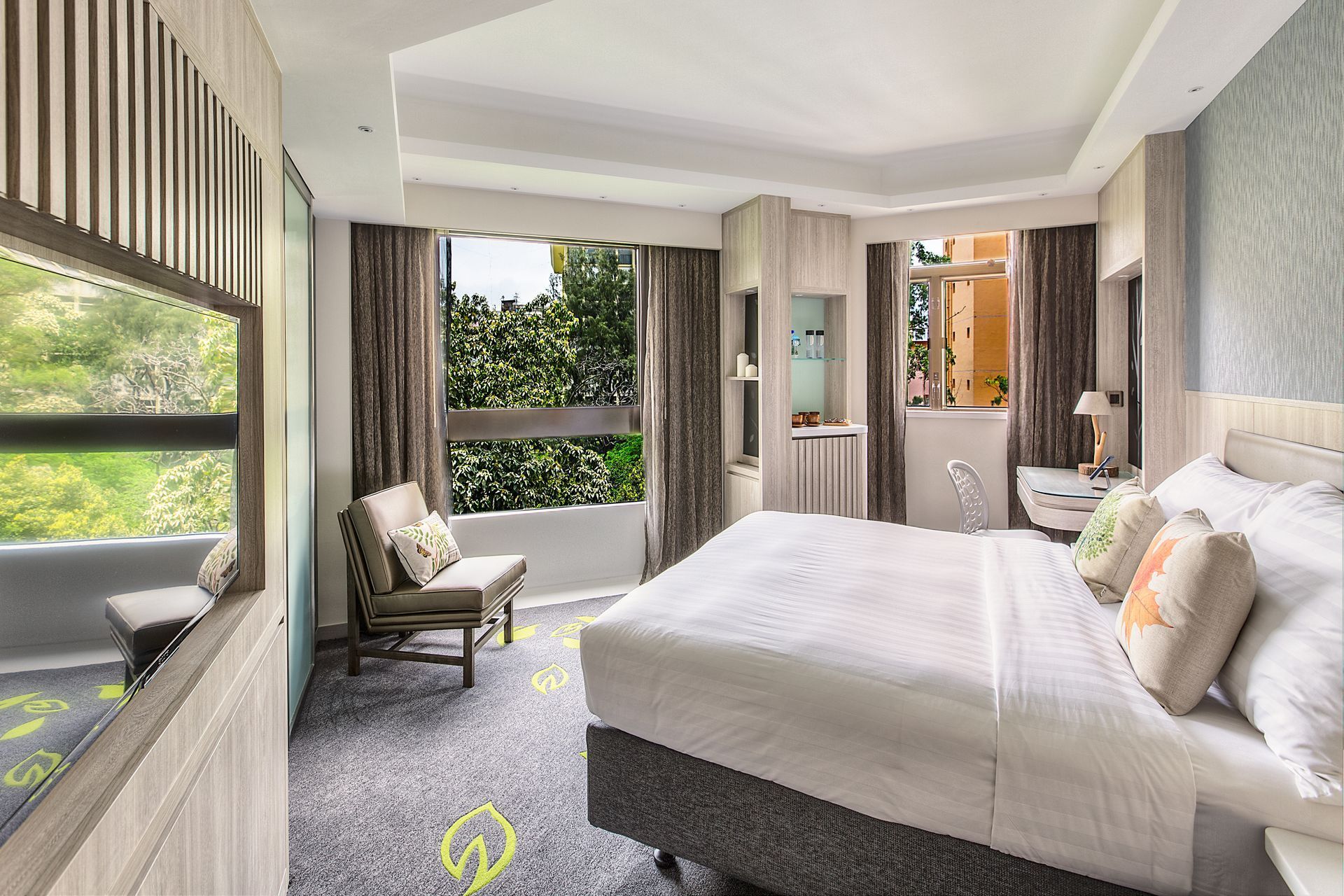 Hotel room with a king-sized bed, armchair, and large windows overlooking trees, muted color scheme.